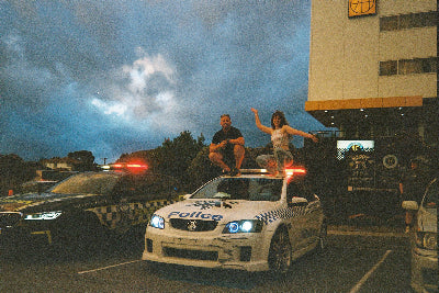 Two people posing on top of a white police car with flashing red lights in a parking lot at dusk, with another police car and a building visible in the background.