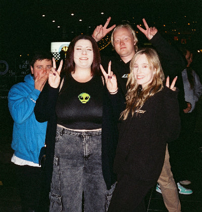 Four young people posing together at a fat ass car meet, making peace signs. The central person wears a black cropped t-shirt with a green alien graphic, wide-leg gray denim cargo pants, and black sneakers.