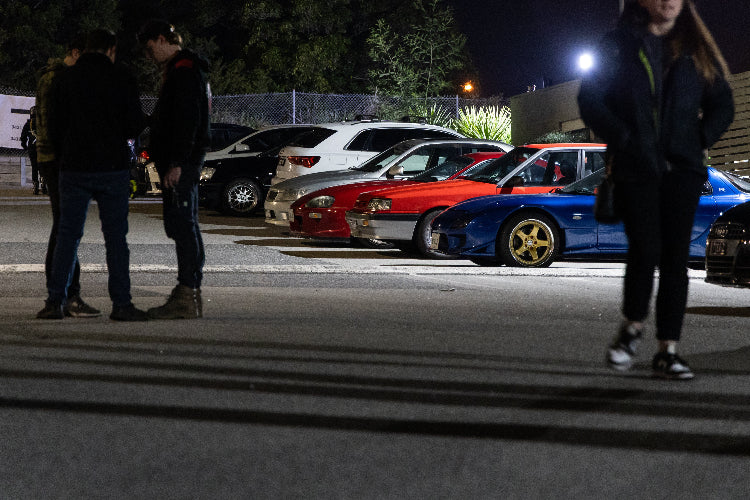 A nighttime car photo at a fat ass car meet parked cars, including a blue car with gold wheels and a red car beside it, with people standing and walking in the foreground.